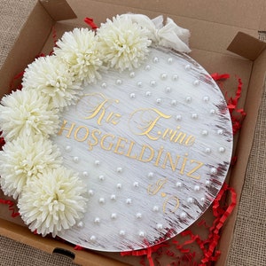 May include: A round, white wooden sign with gold lettering and pearl accents. The sign is decorated with white flowers and is packaged in a cardboard box with red paper filler. The text on the sign reads "Kız Evine Hoşgeldiniz".