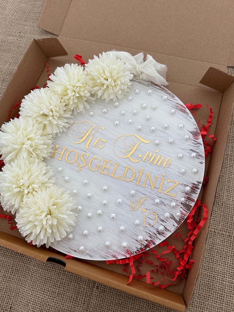 May include: A round, white wooden sign with gold lettering and pearl accents. The sign is decorated with white flowers and is packaged in a cardboard box with red paper filler. The text on the sign reads "Kız Evine Hoşgeldiniz".