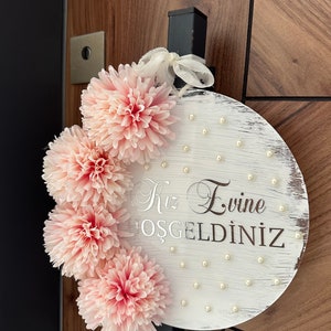 May include: A round, white wooden sign with the text "Kız Evine HOŞGELDİNİZ" in silver script. The sign is decorated with pink fabric flowers and small pearl accents. A white ribbon is attached for hanging.