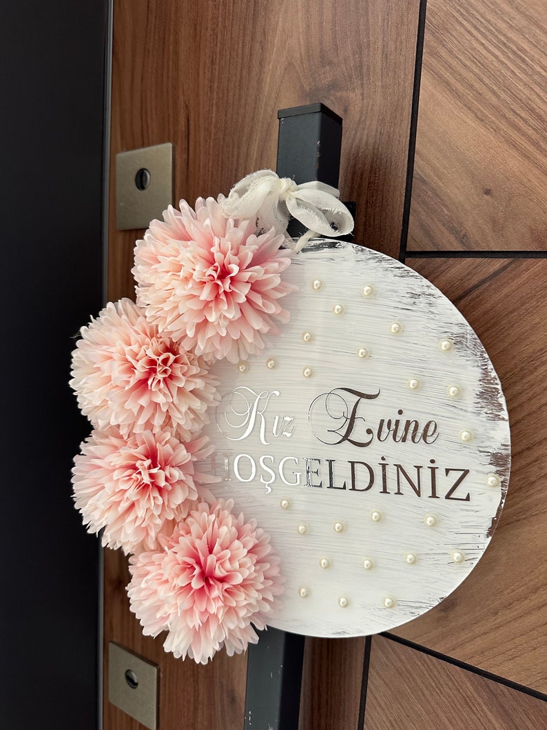 May include: A round, white wooden sign with the text "Kız Evine HOŞGELDİNİZ" in silver script. The sign is decorated with pink fabric flowers and small pearl accents. A white ribbon is attached for hanging.