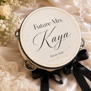 May include: A round tambourine with a cream-colored head featuring the black text Future Mrs. Kaya and Ala&ccedil;atı 2026 with small gold star accents. The instrument has a black rim, silver metal jingles, and is decorated with black satin ribbons.