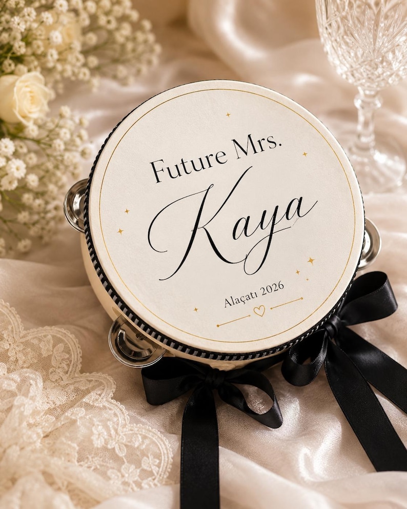 May include: A round tambourine with a cream-colored head featuring the black text Future Mrs. Kaya and Ala&ccedil;atı 2026 with small gold star accents. The instrument has a black rim, silver metal jingles, and is decorated with black satin ribbons.