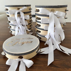 May include: A collection of round wooden tambourines with silver metal jingles. Each tambourine features a black and white striped ribbon trim around the frame and a white satin ribbon bow. One tambourine displays the gold text Damadın Kızı Feyza.