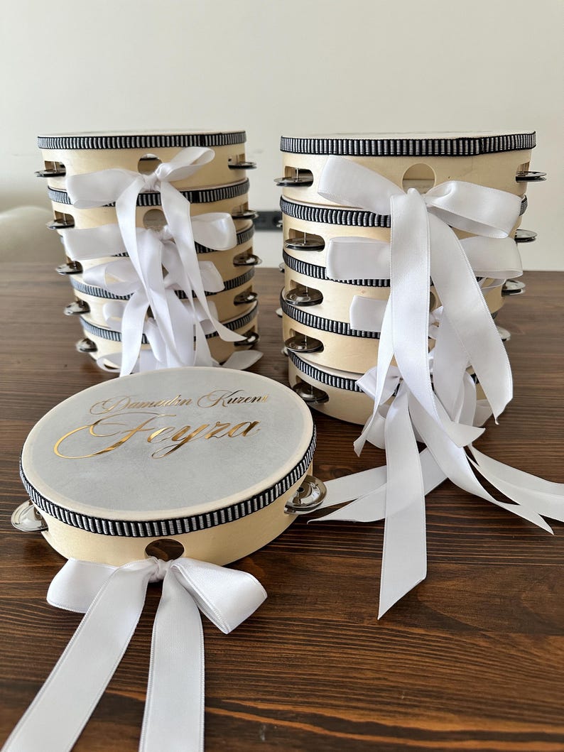 May include: A collection of round wooden tambourines with silver metal jingles. Each tambourine features a black and white striped ribbon trim around the frame and a white satin ribbon bow. One tambourine displays the gold text Damadın Kızı Feyza.