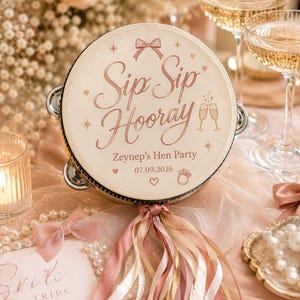 May include: A round tambourine featuring the text "Sip Sip Hooray", "Zeynep's Hen Party", and the date "07.09.2026" in rose gold lettering. The design includes a bow, champagne glasses, hearts, and a ring icon, finished with hanging ribbons.