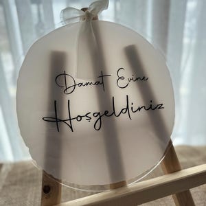 May include: A round, frosted acrylic sign with the words "Damat Evine Hoşgeldiniz" in black script. The sign is attached to a small wooden easel and adorned with a white ribbon. The sign is a wedding or housewarming decoration.