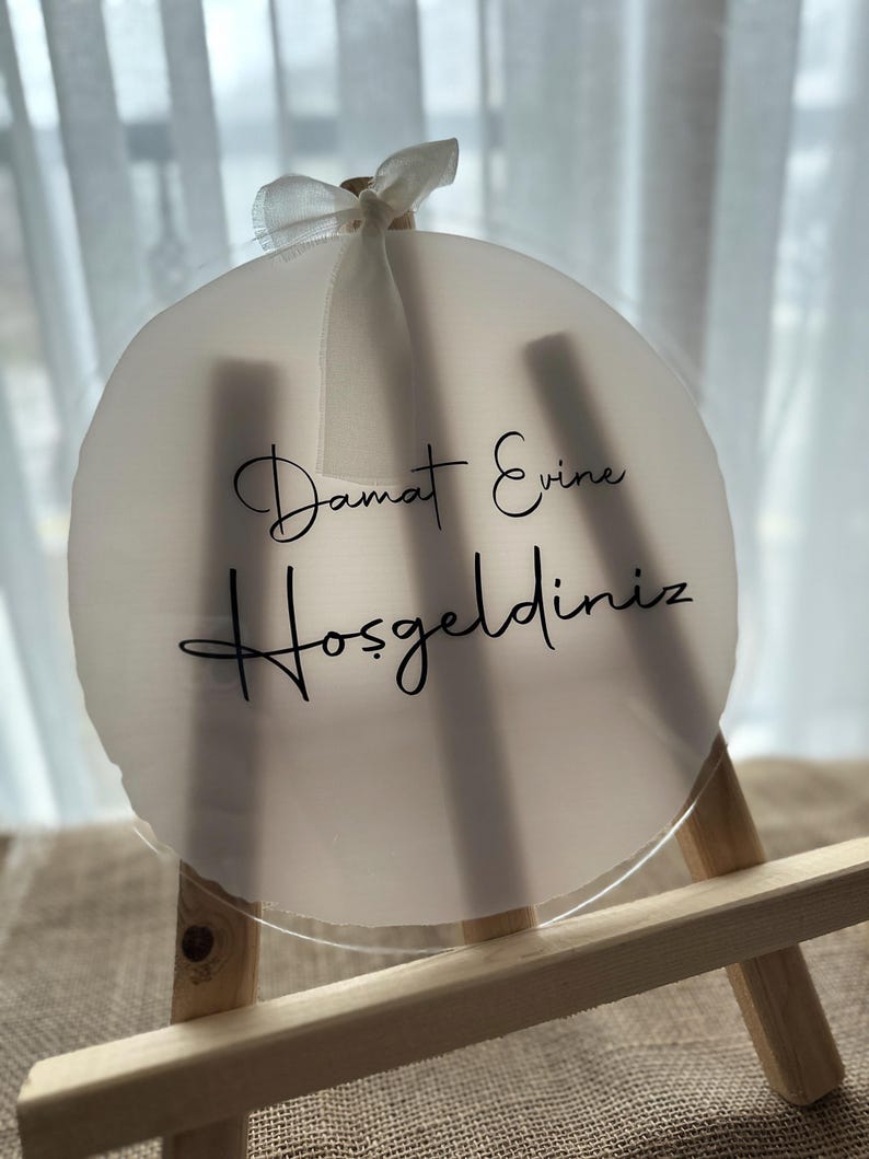 May include: A round, frosted acrylic sign with the words "Damat Evine Hoşgeldiniz" in black script. The sign is attached to a small wooden easel and adorned with a white ribbon. The sign is a wedding or housewarming decoration.