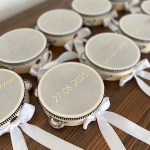 May include: A collection of round tambourines with light beige frames and silver-toned tops. Each tambourine features a black and white striped trim, a white satin ribbon tied in a bow, and gold text including the date 27.08.2025.