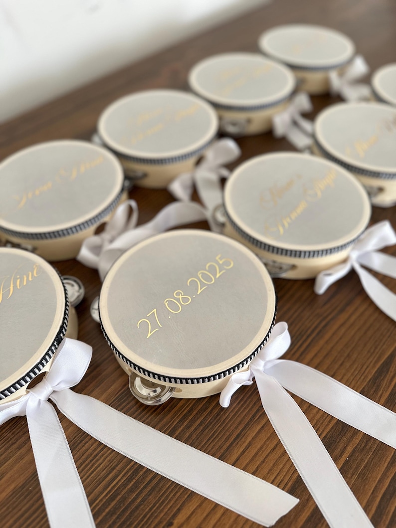 May include: A collection of round tambourines with light beige frames and silver-toned tops. Each tambourine features a black and white striped trim, a white satin ribbon tied in a bow, and gold text including the date 27.08.2025.