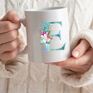 以下が含まれることがあります： 白いセラミック製のマグカップ。青い水彩のEの文字が描かれており、ピンク、青、白の花で飾られています。
