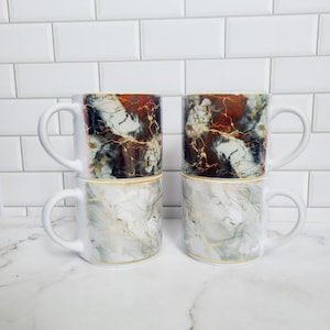 May include: Set of four white ceramic mugs with a marble pattern in shades of grey, black, and gold. The mugs have a gold rim and a white interior.