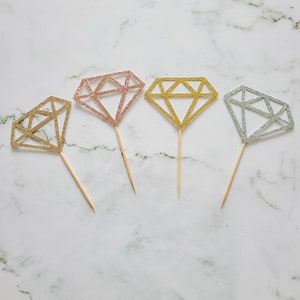 May include: Four diamond-shaped cake toppers in gold, silver, and rose gold glitter. Each topper is attached to a wooden stick.