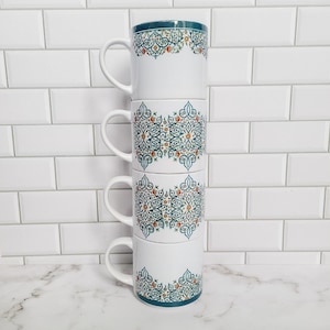 Könnte beinhalten: Ein Stapel von vier weißen Keramikbechern mit einem geometrischen Muster in Teal und Orange. Die Becher sind übereinander gestapelt, wobei der oberste Becher der größte und der unterste Becher der kleinste ist.