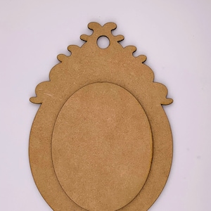 Op de afbeelding: Een ovaalvormig houten frame met een decoratief, versierd ontwerp. Het frame is onbehandeld en klaar om te worden geschilderd of gebeitst.