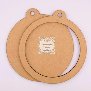 May include: Two circular light brown wooden ornaments with a smooth finish. Each piece features a small tab at the top with a circular hole for hanging. A square sticker in the center displays the text Handcrafted Holiday Traditions.