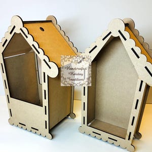 May include: Two unfinished wooden house-shaped boxes. The boxes have scalloped edges and a roof with a contrasting tan interior. The boxes are designed with cut-out details and are suitable for crafting or decorative purposes. The boxes are labeled "Handcrafted Holiday Traditions".