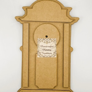 Könnte beinhalten: Ein holzfarbener, uhrförmiger Bastelrahmen mit dekorativer Ober- und Unterseite. Die Uhr hat einen kreisförmigen Ausschnitt und einen rechteckigen Schlitz. Ein Etikett mit der Aufschrift "Handcrafted Holiday Traditions" ist angebracht.