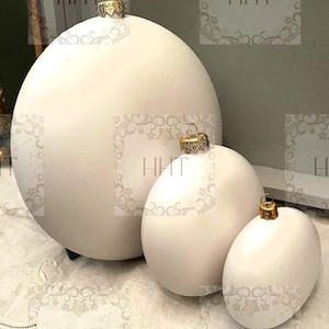 Puede incluir: Tres adornos navideños redondos, blancos y mate con tapas doradas. Los adornos están dispuestos en orden decreciente de tamaño de izquierda a derecha.