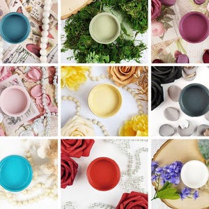 Puede incluir: Nueve pequeños recipientes de pintura abiertos en varios colores, incluyendo turquesa, rosa, amarillo y rojo. Los recipientes están dispuestos sobre superficies con elementos decorativos como flores, cuentas y papel. Los colores son vibrantes.