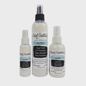 May include: Three white spray bottles of Paint Couture acrylic topcoat gloss. The bottles vary in size, with labels that include "Clear Finish", "UV Protection", and "Water Resistant". The largest bottle contains 237 ml.