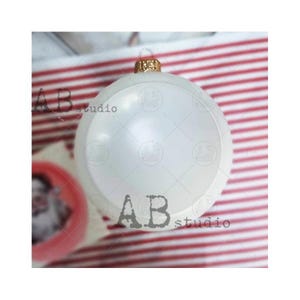 May include: A white, round, plastic Christmas ornament with a gold top. The ornament is sitting on a red and white striped surface.