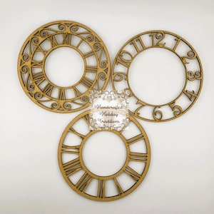 Può includere: Tre quadranti di orologio in legno con numeri romani e arabi. I quadranti sono tagliati in legno marrone chiaro e presentano disegni intricati. Il testo "Handcrafted Holiday Traditions" è al centro.