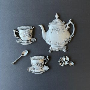 May include: A silver tea set wall decor with a teapot, two cups and saucers, a spoon, and a flower. The set is in a vintage style with ornate details.