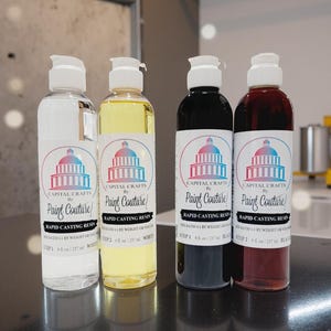 May include: Four bottles of Capital Crafts Paint Couture rapid casting resin. The bottles are clear, yellow, black, and red. Each bottle has a white cap and a label with a Capitol building design and the text "Paint Couture". The bottles are 237 ml.