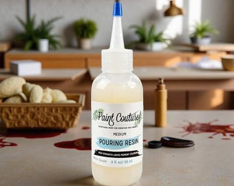 May include: A clear plastic bottle of Paint Couture pouring resin with a white label and blue cap. The label reads "POURING RESIN" and "4 fl oz / 118 ml". The liquid inside is a pale yellow color. The bottle is on a light-colored surface.