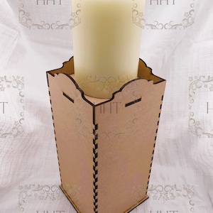 May include: A wooden candle holder with a decorative design. The holder is made of natural wood and has a square base. A white pillar candle is placed inside the holder.