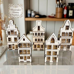 May include: A collection of handcrafted wooden house ornaments with intricate window and door details. The houses are in various architectural styles, with a light wood finish. The image includes a sign that says "Handcrafted Holiday Traditions".