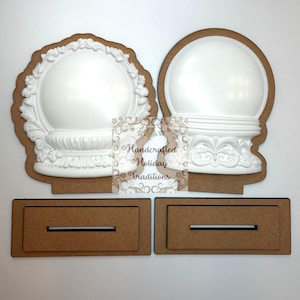 May include: Two white, ornate snow globe bases with decorative details, set on brown wooden cutouts. The bases are accompanied by two rectangular wooden stands with a slot. The image includes the text "Handcrafted Holiday Traditions".