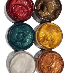May include: Six open jars of metallic paint in various colors, including red, gold, teal, and bronze. The paint has a textured, shimmering appearance, and the jars are arranged in two rows of three.