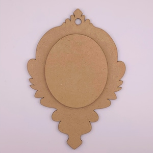 Op de afbeelding: Een onafgewerkt, versierd houten fotolijst met een gat bovenaan om op te hangen. De lijst heeft een decoratieve, ovale uitsparing in het midden.