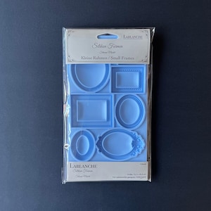 May include: A light blue silicone mold for crafting small frames. The mold, packaged in clear plastic, features various frame shapes, including oval, rectangular, and square designs. The package includes the text "Silikon Formen Kleine Rahmen / Small Frames".