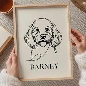 May include: A framed print with a minimalist line drawing of a dog's face, with the name "BARNEY" below. The artwork is in a light wooden frame. The image is held by a person wearing a white sweater.