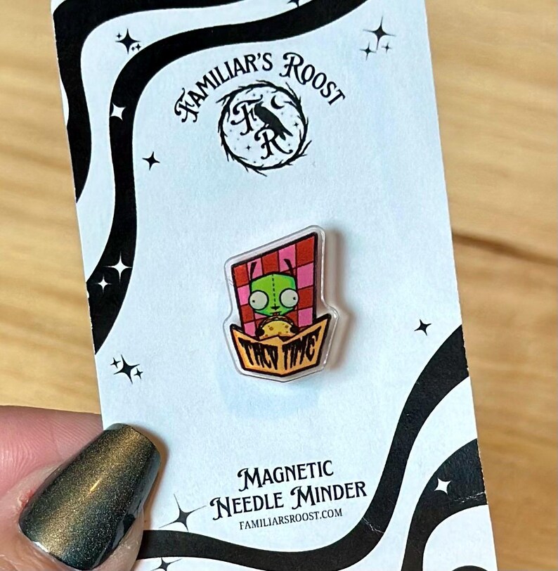 May include: A magnetic needle minder featuring a cartoon character in a taco. The character is green with large eyes, set against a red and pink checkered background. The words "TACO TIME" are visible.