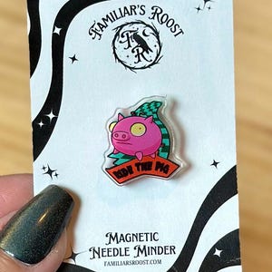 May include: A pink pig-shaped magnetic needle minder with the text "RIDE THE PIG" on a red banner. The minder is on a white card with the text "FAMILIAR'S ROOST" and "MAGNETIC NEEDLE MINDER".