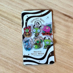 May include: A set of seven colorful magnetic needle minders on a black and white patterned card. The minders feature cartoon characters and phrases like "Ride the Pig" and "Hello World". The card also has the text "Familiar's Roost" and "Magnetic Needle Minder".