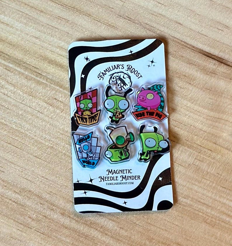 May include: A set of seven colorful magnetic needle minders on a black and white patterned card. The minders feature cartoon characters and phrases like "Ride the Pig" and "Hello World". The card also has the text "Familiar's Roost" and "Magnetic Needle Minder".