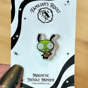 May include: A green cartoon character pin holding pizza, on a white card with black wavy lines and stars. The card reads "Familiar's Roost" and "Magnetic Needle Minder". The pin is a needle minder.