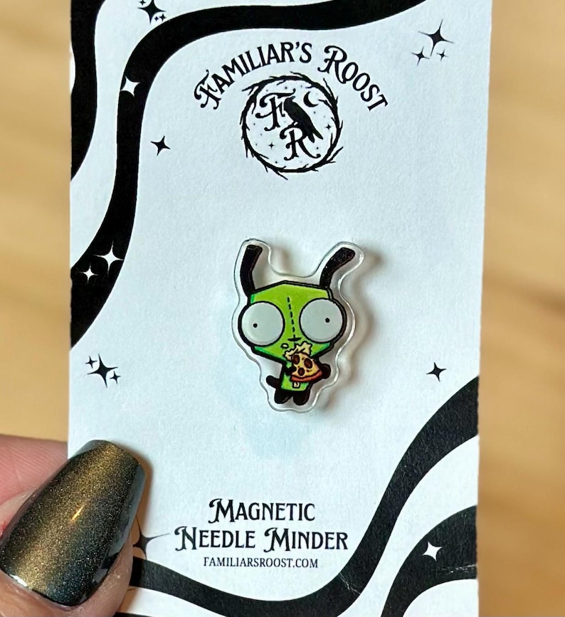 May include: A green cartoon character pin holding pizza, on a white card with black wavy lines and stars. The card reads "Familiar's Roost" and "Magnetic Needle Minder". The pin is a needle minder.