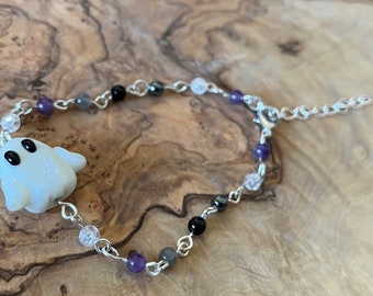 Può includere: Un bracciale a catena color argento con un ciondolo a forma di fantasma bianco con occhi neri. Il bracciale è decorato con piccole perline rotonde nei toni del viola, nero, grigio e trasparente, e include una chiusura a moschettone e una catenella.