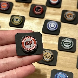 May include: A collection of circular pins featuring various NFL team logos, including the Tampa Bay Buccaneers. The pins are set on small, square black foam pads. The Tampa Bay Buccaneers pin has a red and white design with the team name and logo.