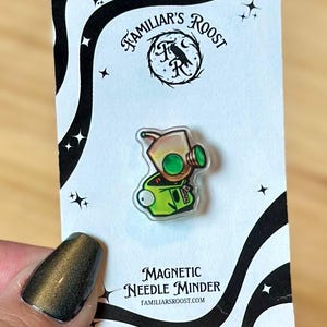 May include: A clear, magnetic needle minder featuring a cartoon character with a tan head, green eyes, and a green outfit. The needle minder is on a white card with black accents and the text "Familiar's Roost" and "Magnetic Needle Minder".