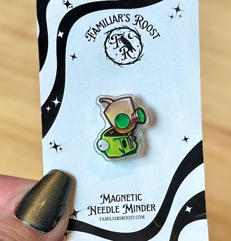 May include: A clear, magnetic needle minder featuring a cartoon character with a tan head, green eyes, and a green outfit. The needle minder is on a white card with black accents and the text "Familiar's Roost" and "Magnetic Needle Minder".