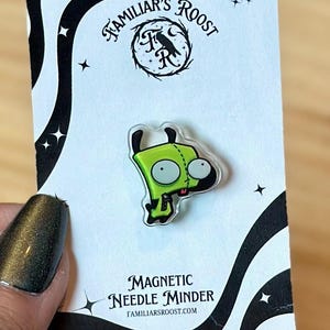 May include: A green cartoon character magnetic needle minder. The minder is on a white card with the text "Familiar's Roost" and "Magnetic Needle Minder". The character has large eyes and a small mouth. The card has a black border with star accents.
