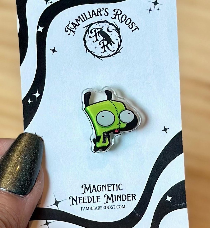 May include: A green cartoon character magnetic needle minder. The minder is on a white card with the text "Familiar's Roost" and "Magnetic Needle Minder". The character has large eyes and a small mouth. The card has a black border with star accents.