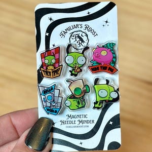 May include: A set of six magnetic needle minders featuring cartoon characters. The minders have various designs, including a green alien with the text "TACO TIME" and a pink pig with the text "RIDE THE PIG". The card reads "FAMILIAR'S ROOST".