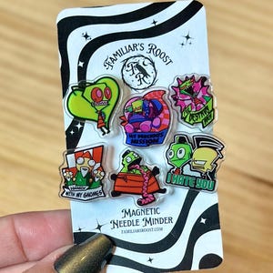 May include: A set of six magnetic needle minders featuring cartoon characters in various poses and colors. The needle minders are on a black and white patterned backing card with the text "FAMILIAR'S ROOST" and "MAGNETIC NEEDLE MINDER".